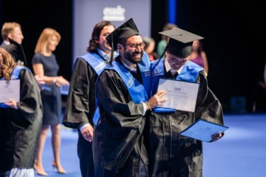 Studierende bei der Exmatrikulation, gekleidet mit Talar und Graduiertenhut. Beide halten eine Urkunde in der Hand