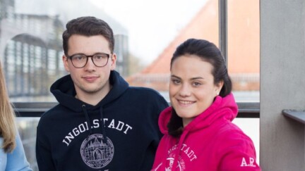 Drei Studierende in Pullovern der Universität vor einem Fenster.