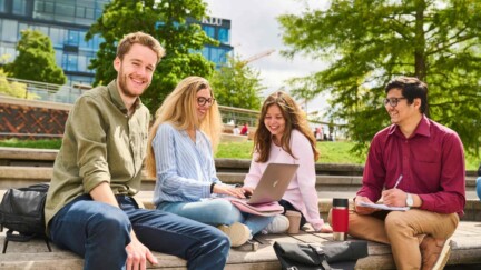 Studierende sitzen auf dem Campus der KLU. Eine Studentin hat einen Laptop vor sich. Ein Student lecht in die Kamera.