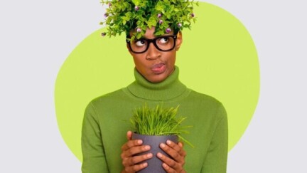 Eine Frau im grünen Pullover hält einen Blumentopf mit Gras. Auf ihrem Kopf befinden sich Zweige eines Laubgewächses. Ihr Gesichtsausdruck ist fragend/nachdenklich.