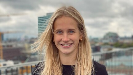 Junge Frau in schwarzem Blazer und Jeans auf einer Dachterrasse, im Hintergrund ein modernes Hochhaus.