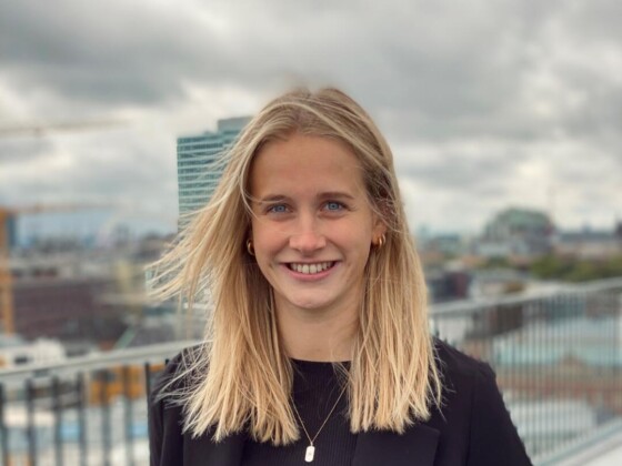 Junge Frau in schwarzem Blazer und Jeans auf einer Dachterrasse, im Hintergrund ein modernes Hochhaus.