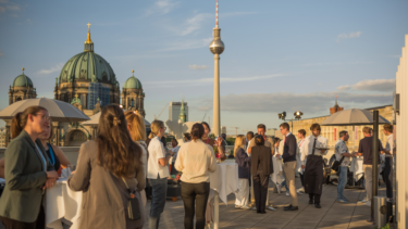 Junge Menschen in Business-Kleidung stehen in Grüppchen zusammen und unterhalten sich. Sie stehen auf einer Dachterrasse, hinter ihnen der Berliner Fernsehturm.