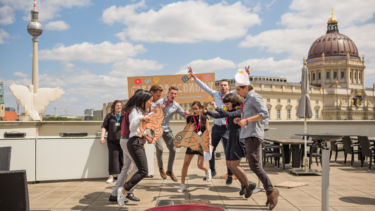 Junge Menschen, die gemeinsam auf einer Dachterrasse in die Höhe springen, hinter ihnen der Berliner Fernsehturm.