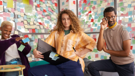 Zwei Frauen und ein Mann kniend in einem Büro, auf der Glaswand hinter ihnen viele bunte Post-Its.