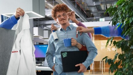 Mann steht mit Laptop in einem Büro. Ihm werden von anderen Mitarbeitern verschiedene Dinge gereicht.