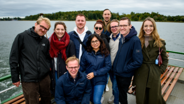 Junge Frauen und Männer in Wind- und Regenjacken stehen auf einem Boot, um sie herum Wasser und im Hintergrund ein bewaldetes Stück.