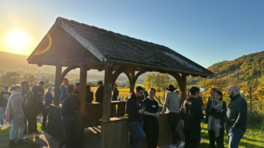 Frauen und Männer stehen in Outdoorkleidung um einen hölzernen Unterstand in einer grünen, hügeligen Landschaft, von links oben fällt Sonnenschein ins Bild.