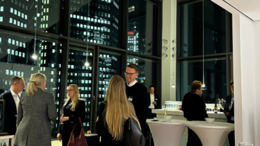 In einem verglasten Bürogebäude mit hohen Wänden unterhalten sich Frauen und Männer in Business-Kleidung, außerhalb des Fensters herrscht Nacht und sind einzelne Fenster von weiteren Bürogebäuden erleuchtet.