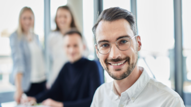 Ein Mann mit Brille und im Hemd lächelt in die Kamera, hinter ihm ein Büro und etwas verschwommen die Schemen von zwei Frauen und einem Mann.