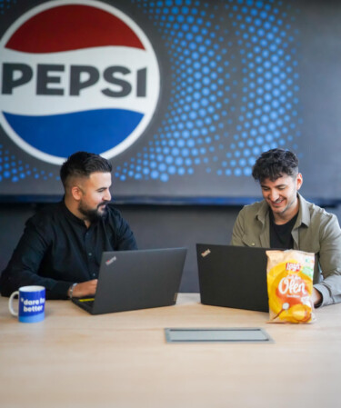 Zwei Mitarbeiter arbeiten an ihren Laptops und sitzen unter dem blau-roten Logo von Pepsi.
