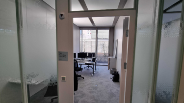 Blick vom Flur in ein langgezogenes Büro mit hellem Boden, in das aus dem Fenster Licht fällt.