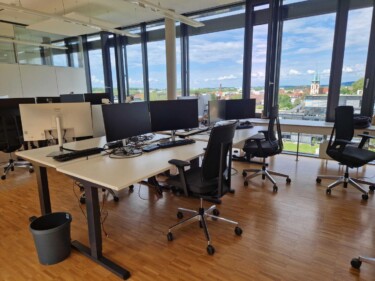 Ein Büro mit Holzboden, mehreren weißen Schreibtischen, Bildschirmen, Bürostühlen, im Hintergrund eine Glasfassade.