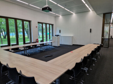 Konferenzraum mit U-förmig angeordneten Tischen, Stühlen, einem Beamer und Blick ins Grüne.