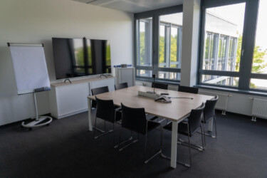 Konferenzraum mit einem Tisch, mehreren Stühlen drumherum, einer Freisprechanlage, dahinter ein Flachbildschirm, daneben ein Whiteboard.