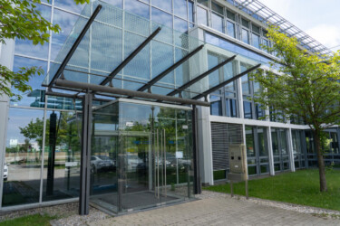 Ein Gebäude mit Glasfassade von außen, auf den Türen das blaue Logo von TNG Technology Consulting, davor ein gepflasterter Weg, links und rechts Wiese.