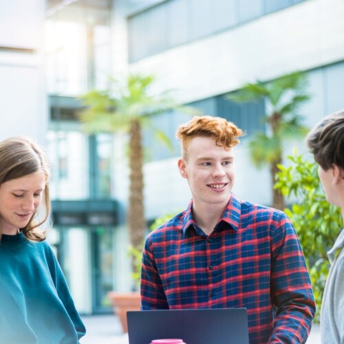 drei Studierende stehen vor einem Universitätsgebäude und unterhalten sich