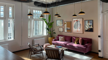 Ein helles Zimmer mit Fenstern, von der Decke hängen mehrere Lampen, an der Wand hängen Bilder, auf dem Boden stehen eine lilafarbene Couch, zwei Sessel und ein kleiner Tisch.