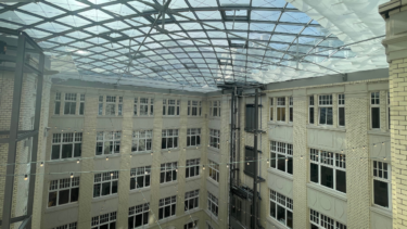 Blick auf ein beigefarbenes, im Viereck gebautes Gebäude, das von einer Glaskuppel überdacht wird.