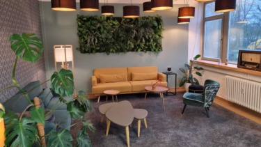 Ein Raum mit einer bunten Wand, mit Teppich, Sofa, Sessel, Beistelltisch, Pflanzen und vielen Lampen